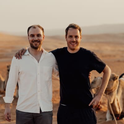 Two men smiling and posing with their arms around each other in a desert landscape with camels in the background.