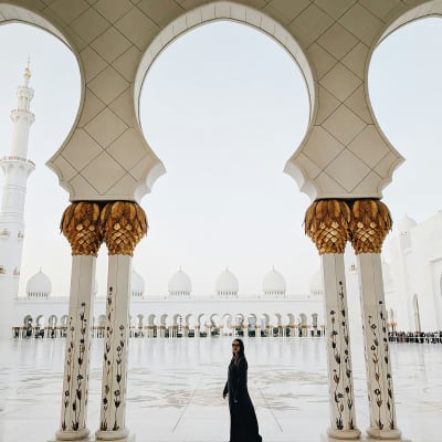 A woman in black abaya standing inside the Sheikh Zayed Grand Mosque with white domes and arches in the background.