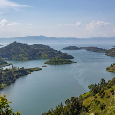 Ein weiser See, umgeben von üppigen grünen Hügeln unter einem wolkenverhangenen Himmel, mit entfernten Bergen im Hintergrund.