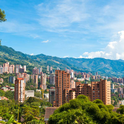 A panoramic view of Medellin, Colombia, with tall residential buildings, lush greenery, and mountain ranges in the background under a clear blue sky.