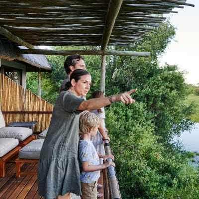 Eine Familie steht auf der Holzveranda einer Lodge und blickt auf die üppige grüne Landschaft und einen Fluss oder See.