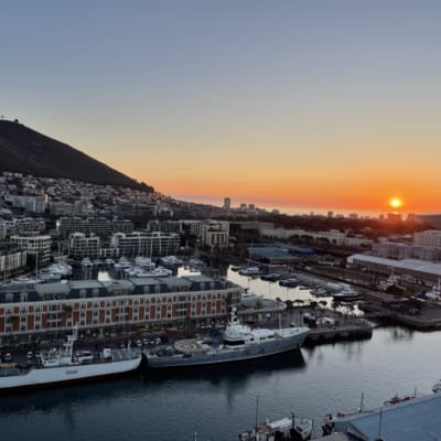 A harbor at sunset with boats docked and a cityscape behind, with a hill on the left and the sun setting on the horizon.