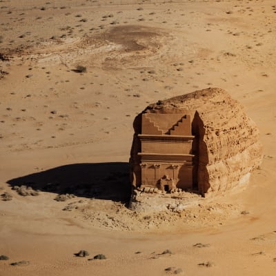 A large ancient tomb carved into a rock in a desert landscape with sandy terrain and some footprints.