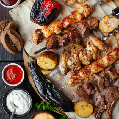 A platter of grilled meats, vegetables, and sauces on parchment paper, with small bowls of condiments and a dark wooden table.
