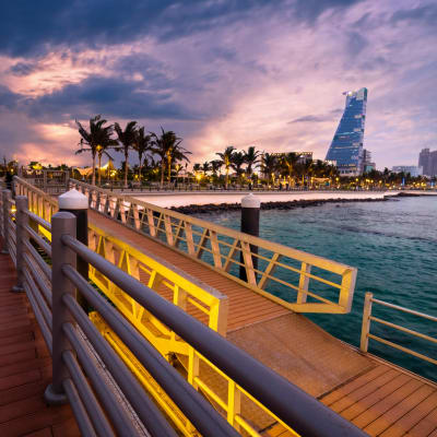 Scenic view of the Jeddah waterfront boardwalk with palm trees and modern buildings in the distance during dusk.