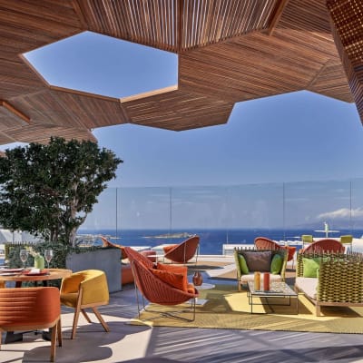 Outdoor terrace with colorful chairs, sofas, and a large tree, overlooking the sea in Greece.
