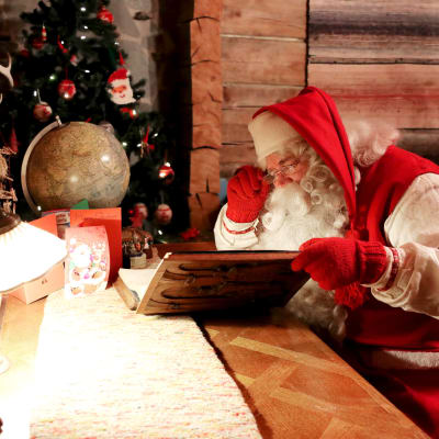 Weihnachtsmann in traditionellem rotem Gewand sitzt an einem hölzernen Tisch in einem rustikalen Raum und betrachtet konzentriert ein aufgeschlagenes Buch oder eine Karte, während im Hintergrund ein weihnachtlich geschmückter Tannenbaum und ein Globus sichtbar sind.