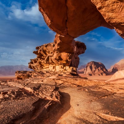 A desert landscape with large, weathered red rock formations under a blue sky with clouds.