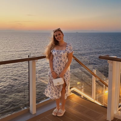 Eine junge Frau in einem geblümten Kleid und Sandalen steht lächelnd auf dem Deck eines Schiffs bei Sonnenuntergang, im Hintergrund sind Meer und entfernte Inseln zu sehen.