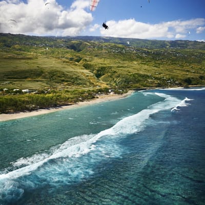 Luftaufnahme einer Küstenlandschaft mit türkisfarbenen Meereswellen, die an einem schmalen Strand brechen, umgeben von grünen Hügeln, unter einem blauen Himmel mit Wolken und mehreren Paragleitern im Flug.