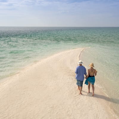 Ein Paar geht Hand in Hand entlang eines Sandpfades an einem Strand, mit dem Ozean im Horizont unter einem teils bewölkten Himmel.