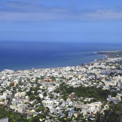 Luftaufnahme der Stadt Saint-Denis entlang der Küste mit blauem Ozean bis zum Horizont unter teilweise bewölktem Himmel.
