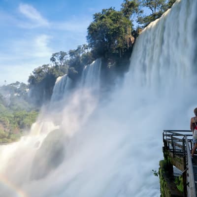Zwei Personen stehen auf einer Aussichtsplattform mit Blick auf die beeindruckenden Iguazu-Wasserfälle, umgeben von üppigem Grün unter einem klaren blauen Himmel.