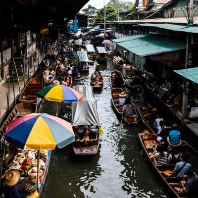 Boote, die durch einen belebten schwimmenden Markt mit Verkäufern und Einkäufern unter bunten Regenschirmen fahren.