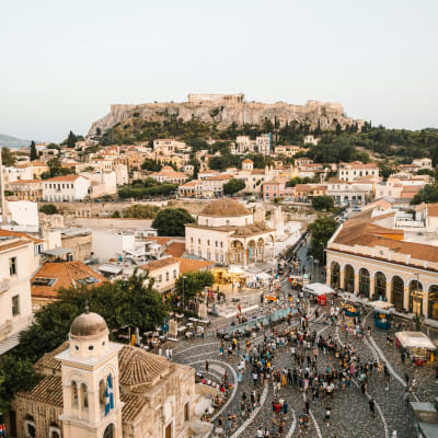 Belebter Fußgängerplatz in Athen mit historischen Gebäuden und dem Akropolis-Hügel im Hintergrund unter klarem Himmel.