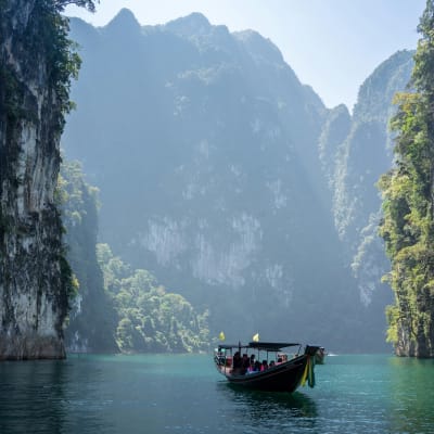A boat sailing on a calm river surrounded by tall, lush green limestone cliffs under a clear sky.