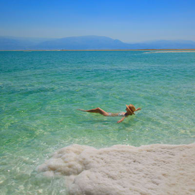 A person floating peacefully on the clear, salty waters of the Dead Sea with a salt-covered shoreline in the foreground.