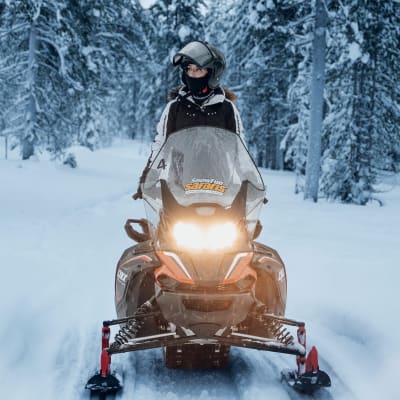 Person wearing a helmet and winter gear riding a snowmobile on a snowy trail in a dense forest covered with snow.