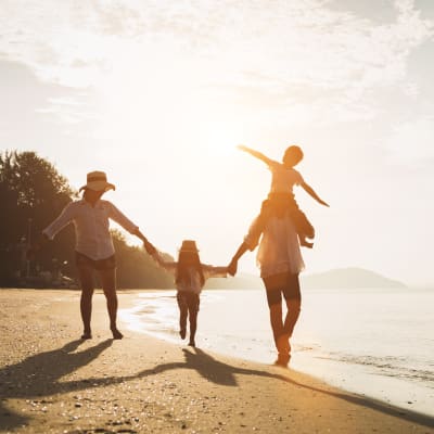 Eine Familie spaziert am Strand bei Sonnenuntergang, ein Kind wird auf den Schultern eines Erwachsenen getragen und ein anderes hält die Hand eines Elternteils.