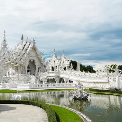 A stunning white temple with ornate architectural details, surrounded by a pond and lush greenery, under a cloudy sky.