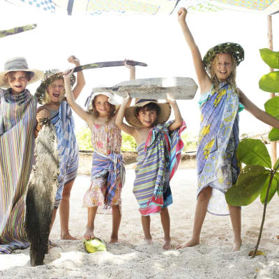 Children playing on the beach with makeshift weapons and a large leaf in a sunny, tropical setting.