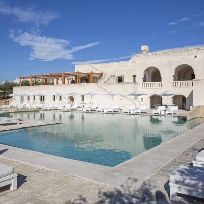 Ein luxuriöser Hotelpoolbereich mit weißen Liegen und einem großen Swimmingpool, vor einem weißen Gebäude mit Bögen und Terrassen unter einem blauen Himmel.
