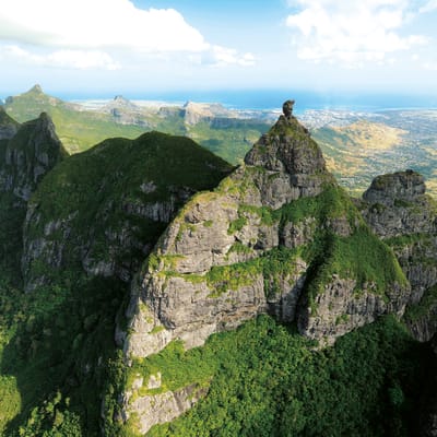 Aerial view of lush green mountains with steep rocky peaks and a distant coastline.