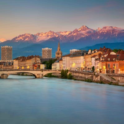 A panoramic view of Grenoble, France, showing a river in the foreground, colorful buildings along the riverbank, and snow-capped mountains in the background during sunset.