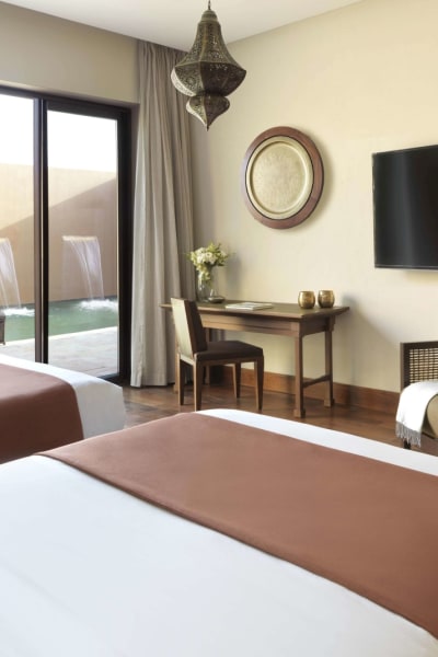 Zwei Einzelbetten mit braunen Bettläufern in einem hellen Hotelzimmer mit Blick auf einen Pool und eine Terrasse durch Schiebetüren.