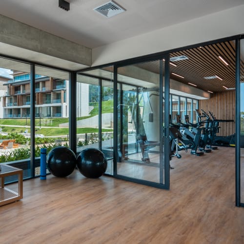 Moderner Fitnessraum mit bodentiefen Fenstern, die auf eine grüne Außenanlage mit Liegestühlen und einem Pool blicken, ausgestattet mit Fahrradergometern und großen Gymnastikbällen.