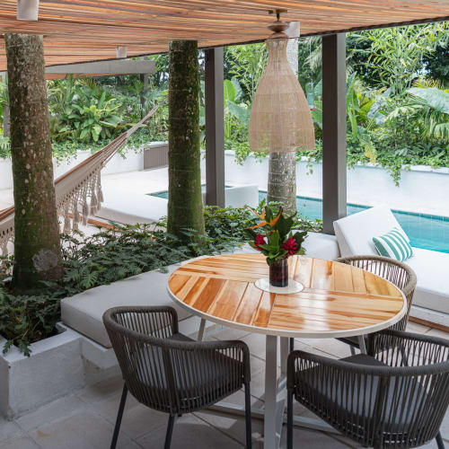 A covered outdoor patio with a wooden ceiling, featuring a person relaxing in a hammock surrounded by tropical plants, a pool in the background, and a round wooden table with chairs in the foreground.
