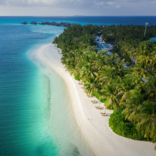 Aerial view of a tropical island beach with white sand, lined with palm trees and lounge chairs, next to clear turquoise water with a small boat floating nearby.