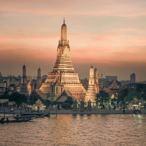 Illuminated Wat Arun temple at dusk along the Chao Phraya River in Bangkok with a long-tail boat in the foreground.