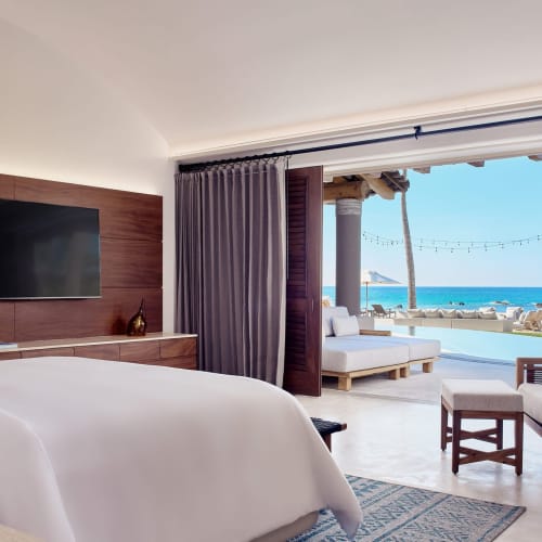 Luxuriöses Schlafzimmer mit großem Bett, Flachbildfernseher und offenem Schiebetür zu einer Terrasse mit Sitzbereich und Blick auf das Meer und Palmen.