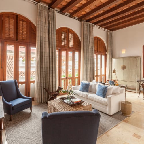 A bright living room with white walls, wooden ceiling beams, and large arched windows covered with beige curtains. The room features a white sofa with blue cushions, two dark blue armchairs, a wicker coffee table with books and flowers, and a small dining table near a wooden cabinet and window.