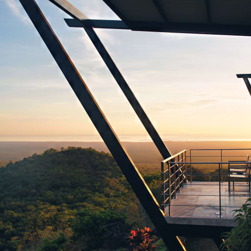 Balkon eines modernen Lodgezimmers mit Blick auf einen bewaldeten Hügel bei Sonnenuntergang, mit einem Stuhl auf dem Balkon und großen schrägen Stützstreben.