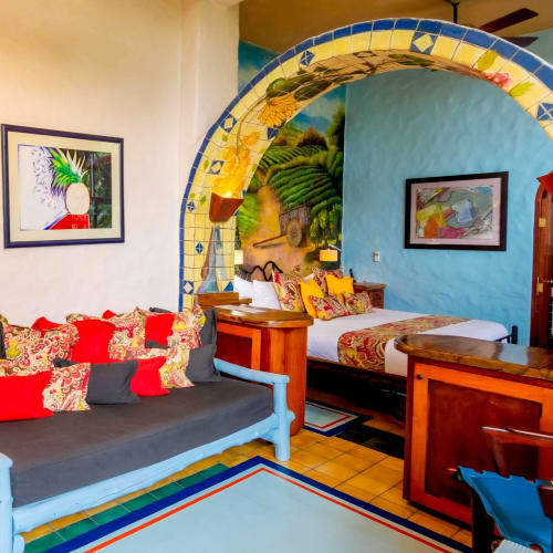 A brightly decorated rustic bedroom with a tiled archway leading to a wooden bed adorned with colorful pillows and throws, a gray couch with vibrant cushions, and tropical-themed artwork on the walls.