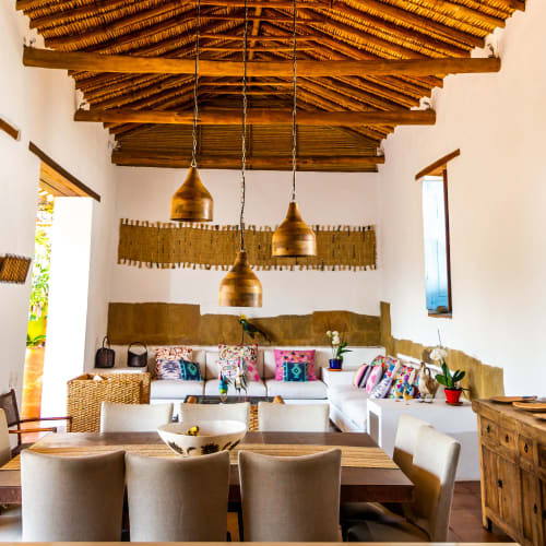 Interior of a rustic living and dining area with a thatched wooden ceiling, hanging pendant lights, a wooden dining table with beige chairs, and a white sofa adorned with colorful cushions and decorative plants.