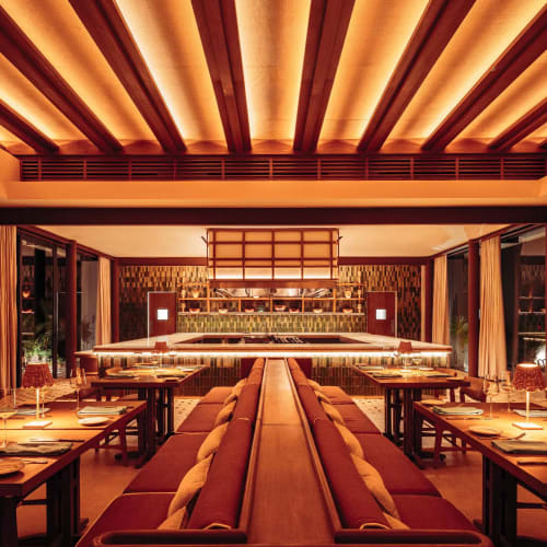 Warmly lit restaurant interior with a central long bench seating and tables arranged along the sides, featuring modern wooden ceiling beams and curtains on windows.