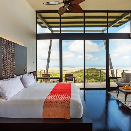 Modernes Hotelzimmer mit einem Kingsize-Bett mit weißen Bettwäsche und orangem Überwurf, großen Glastüren, die sich zu einem Balkon mit Stühlen öffnen, mit Blick auf grüne Hügel und das entfernte Meer unter einem teilweise bewölkten Himmel.