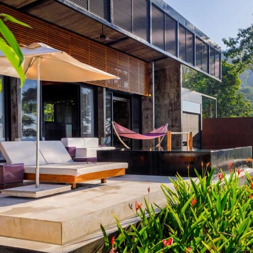 Moderner Außenbereich einer Boutique-Hotel Suite mit Liegestuhl und Sonnenschirm, Hängematte und einem schwarzen rechteckigen Pool, umgeben von Grün und mit Blick auf einen bewaldeten Hügel.