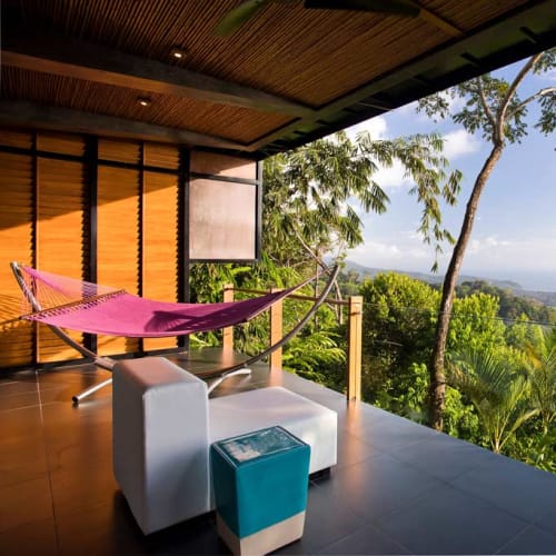 Balkon einer Suite im Kura Boutique Hotel mit einer pinken Hängematte, weißem gepolstertem Sitz, kleinem blauem Tisch und Panoramablick auf üppiges Grün und die entfernte Küste unter klarem Himmel.