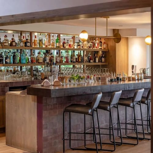 Modern bar interior with high stools lined up along a brick counter and shelves stocked with various bottles and glassware.