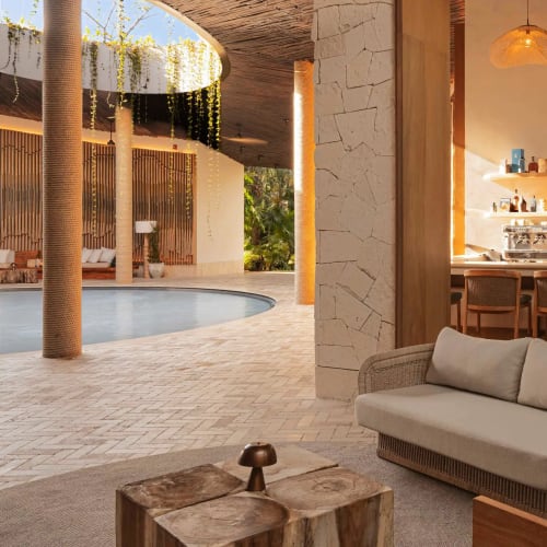 Outdoor lounge area with cushioned wicker sofa and wooden tables near a bar with shelves and chairs, under a shaded roof overlooking a pool.