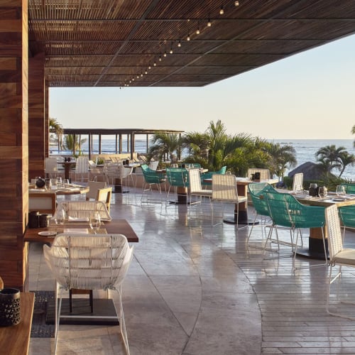 Außenbereich des Restaurants im Four Seasons Resort Punta Mita mit Tischen und Stühlen unter einem schattigen Dach, mit Blick auf Palmen und das Meer bei Sonnenuntergang.
