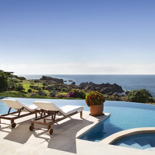 Infinity-Pool mit Liegestühlen mit Blick auf felsige Küste und Meer unter klarem Himmel.
