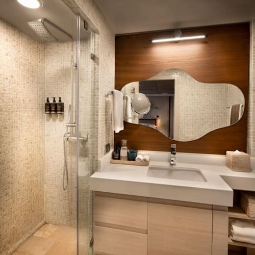 Modernes Badezimmer mit gefliestem Duschbereich, großem unregelmäßig geformten Spiegel und Waschbecken auf weißer Arbeitsplatte mit Holzschränken darunter.