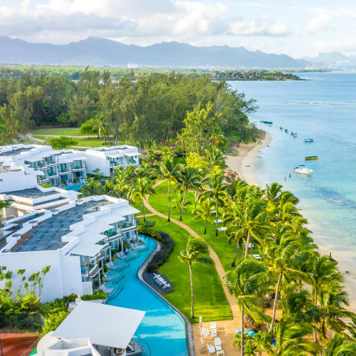 Aerial view of a resort with white buildings and a long swimming pool adjacent to a sandy beach lined with palm trees, overlooking a calm sea and distant mountains under a partly cloudy sky.