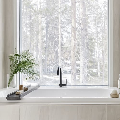 Minimalistic bathroom with a white bathtub positioned in front of a large window showcasing a frosted view of snow-covered trees outside.