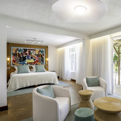 Bright and airy hotel room with a large bed, two white armchairs, a small rattan table and stools, large windows with white curtains, and a sliding glass door opening to a garden with tropical plants.